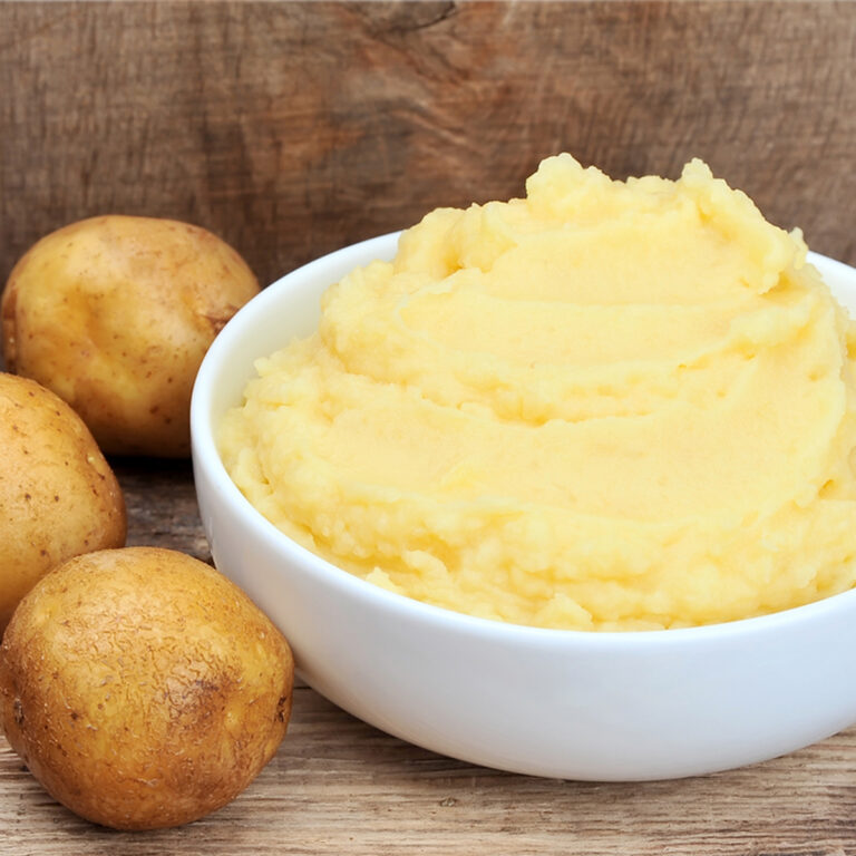 A bowl of mashed potatoes with three whole potatoes sitting beside it.