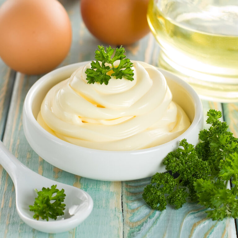 A bowl fo mayonnaise garnished with parsley with a bowl of oil and two eggs in the background