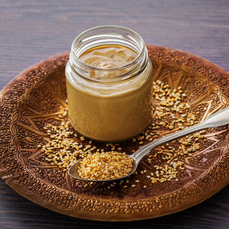 A jar of tahini and a spoonful of sesame seeds sitting on a wooden plate