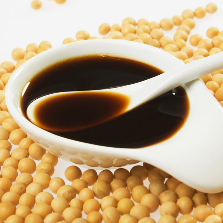 A small dish of tamari with a spoon in it, surrounded by soy beans.