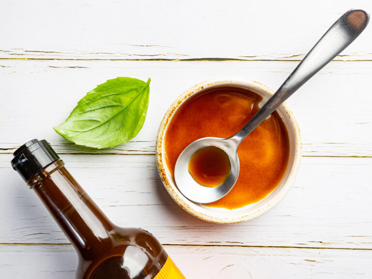 Overhead view of a small bowl of worcestershire sauce with a spoon in it with a bottle and basil leaf beside it.