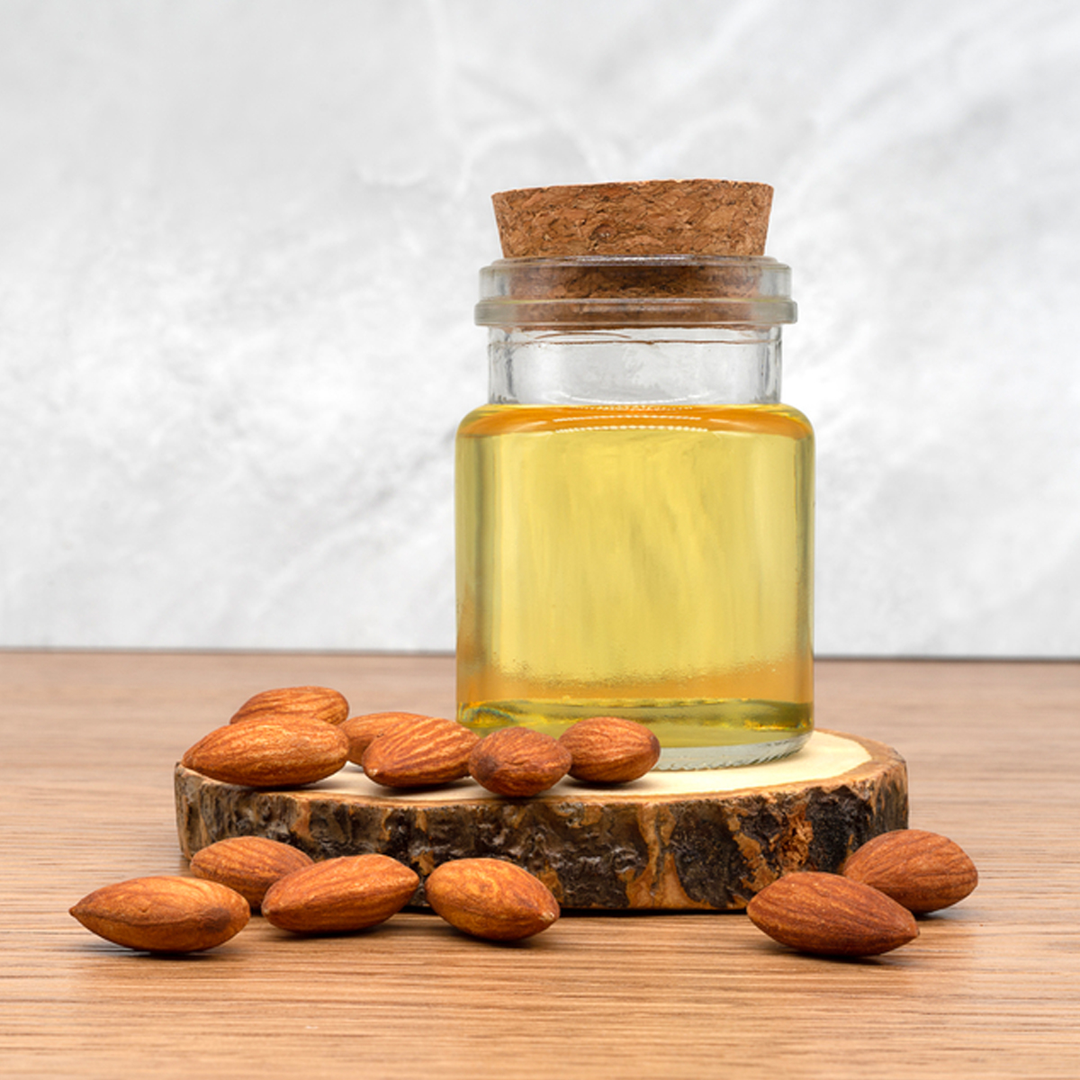A jar of almond oil sitting on a piece of wood surrounded by almonds.