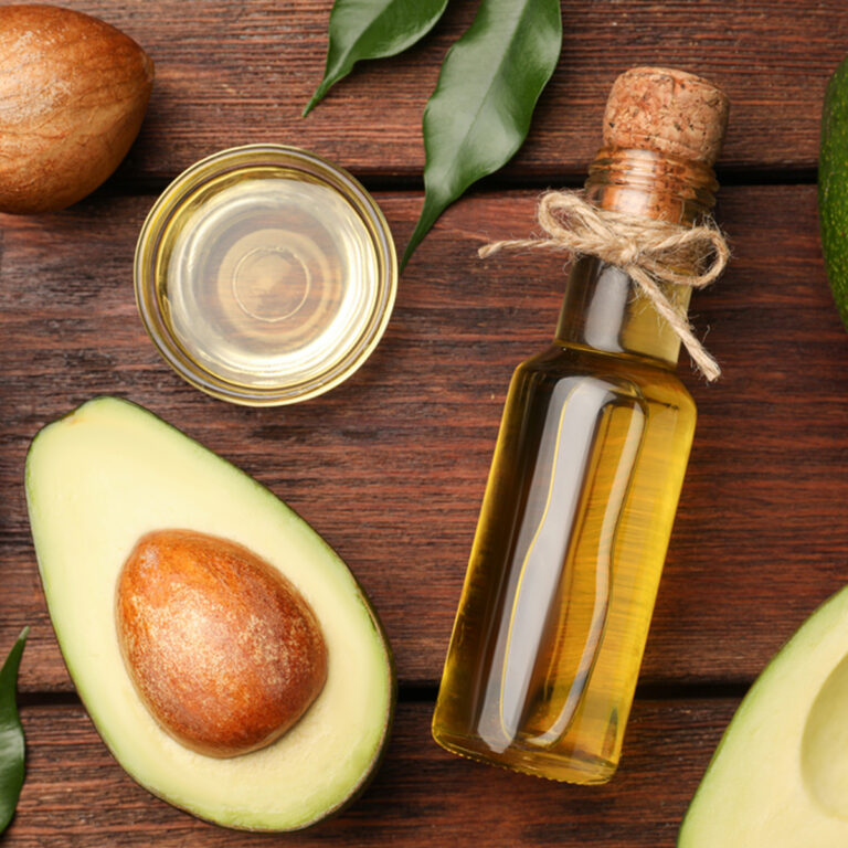 A bottle of avocado oil laying on a wooden tabletop with a small bowl of oil and a halved avocado beside it.