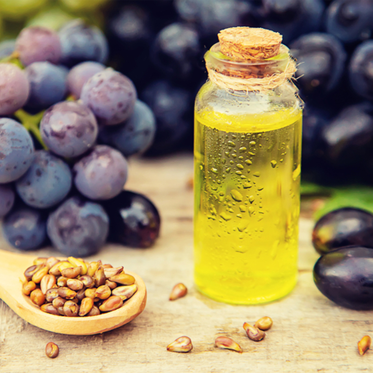 A small jar of grapeseed oil next to a spoonful of grape seeds with a bunch of purple grapes in the background.