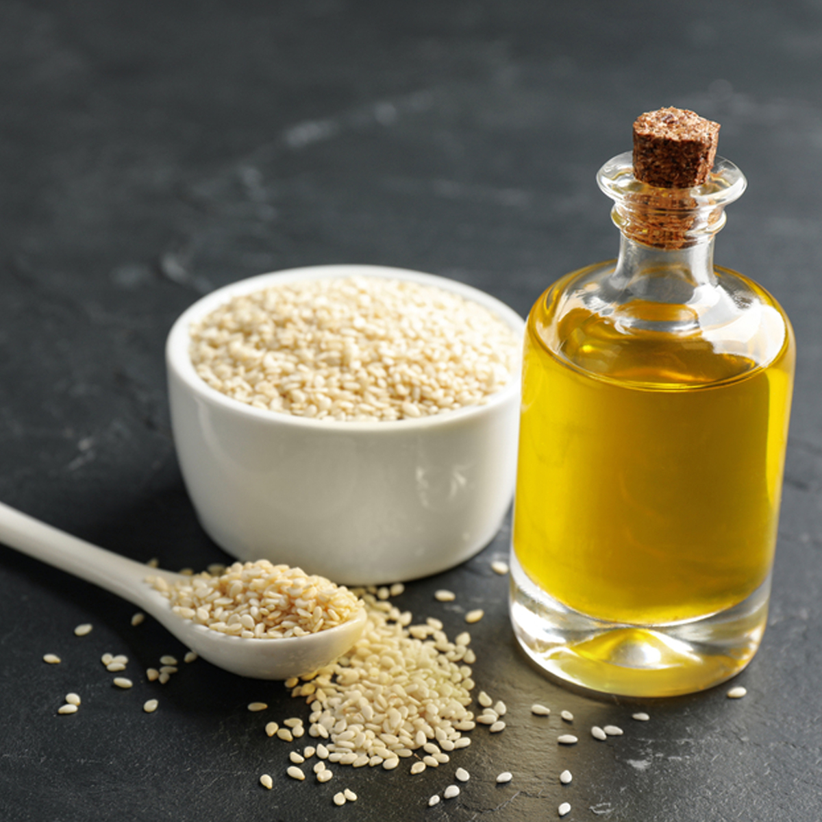 A small bottle of sesame oil next to a bowl of sesame seeds and a spoonful of sesame seeds