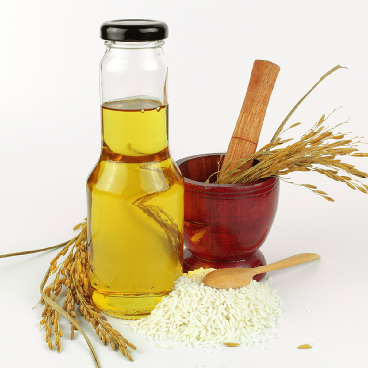 A bottle of rice bran oil sitting beside a pile of rice with a mortar and pestle in the background containing a sprig of dried bran.