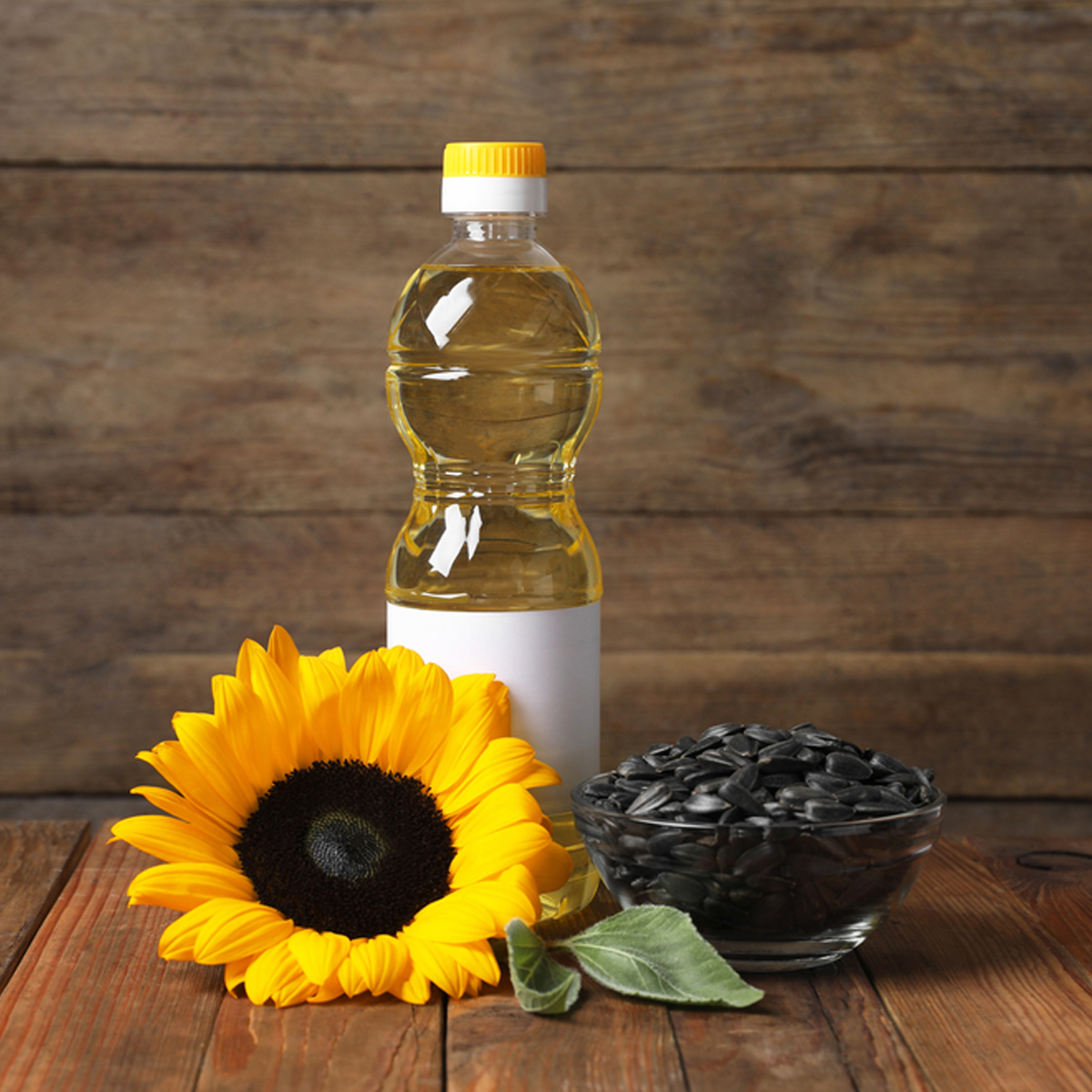 A plastic bottle of sunflower oil with a bowl of sunflower seeds beside it and a sunflower in the foreground.
