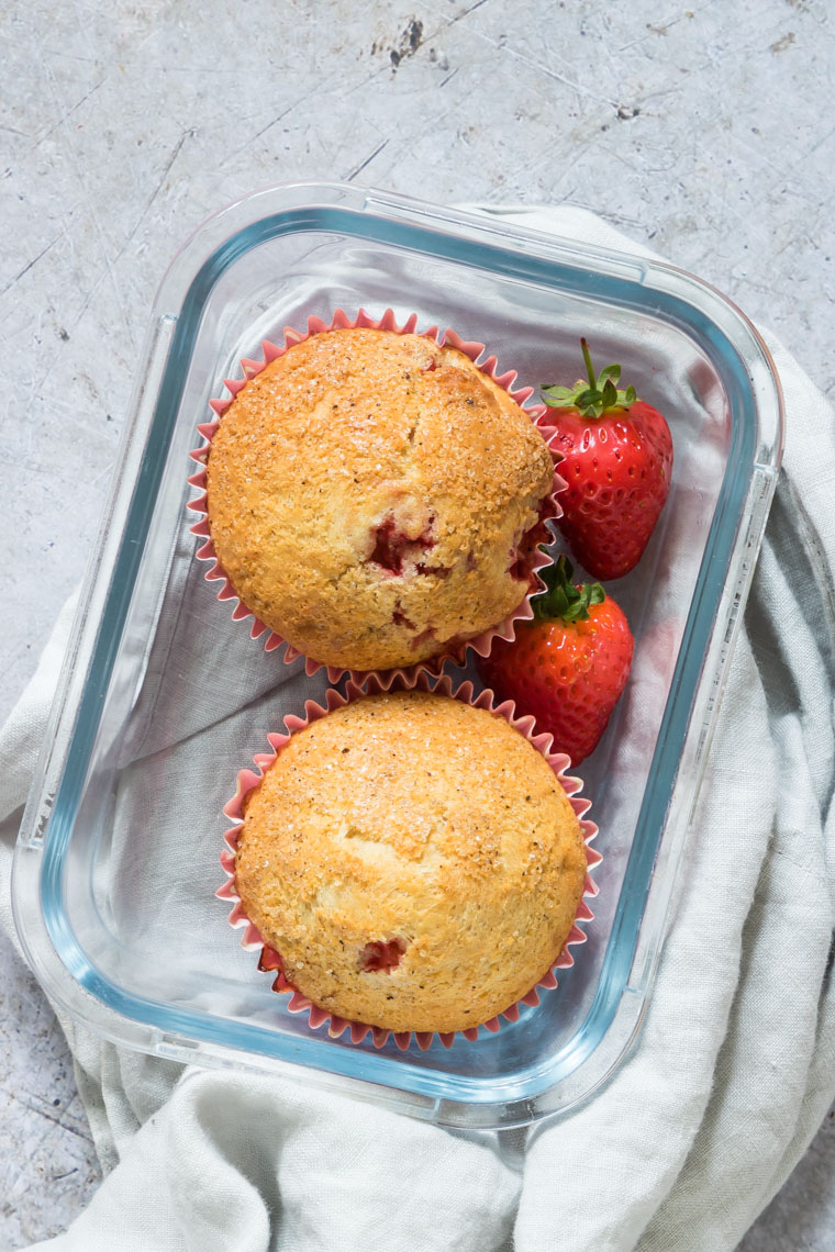 two muffins in a glass meal storage container with two strawberries