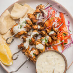 Overhead view of Greek chicken kebabs on a white plate with tzatziki and other sides.