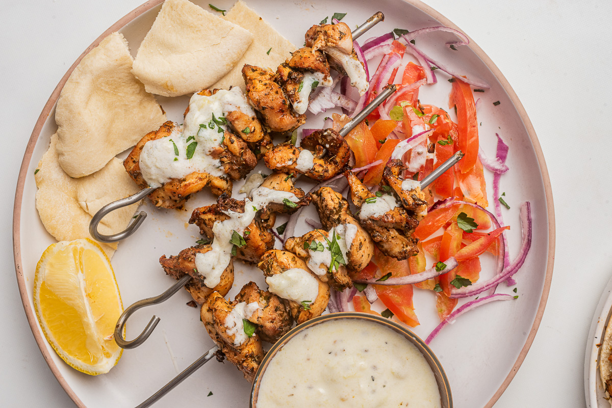 Horizontl overhead view of chicken kabobs on a white plate with tzatziki and other sides.