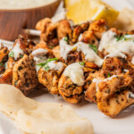 Close up view of skewered chicken on a white plate with tzatziki sauce over them.