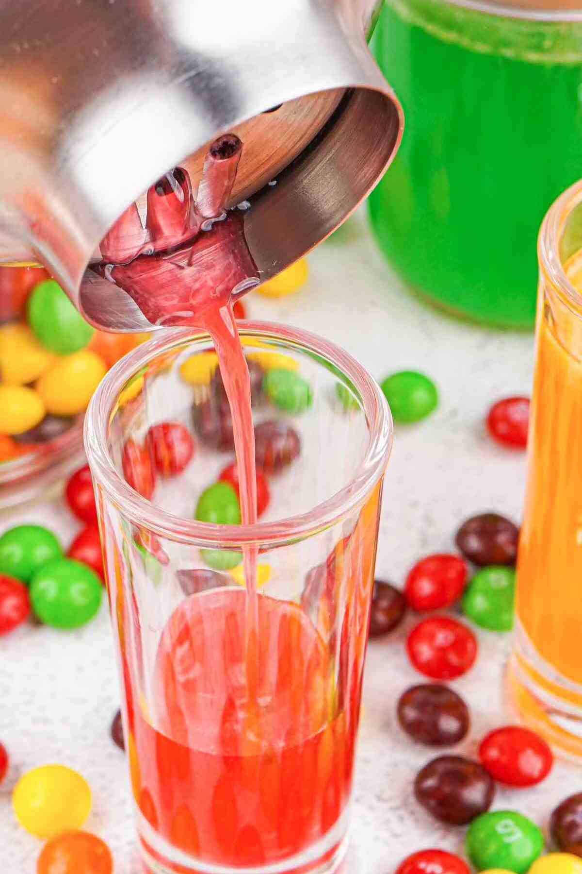 the red version of the skittles shot being poured out of a cocktail shaker into a glass