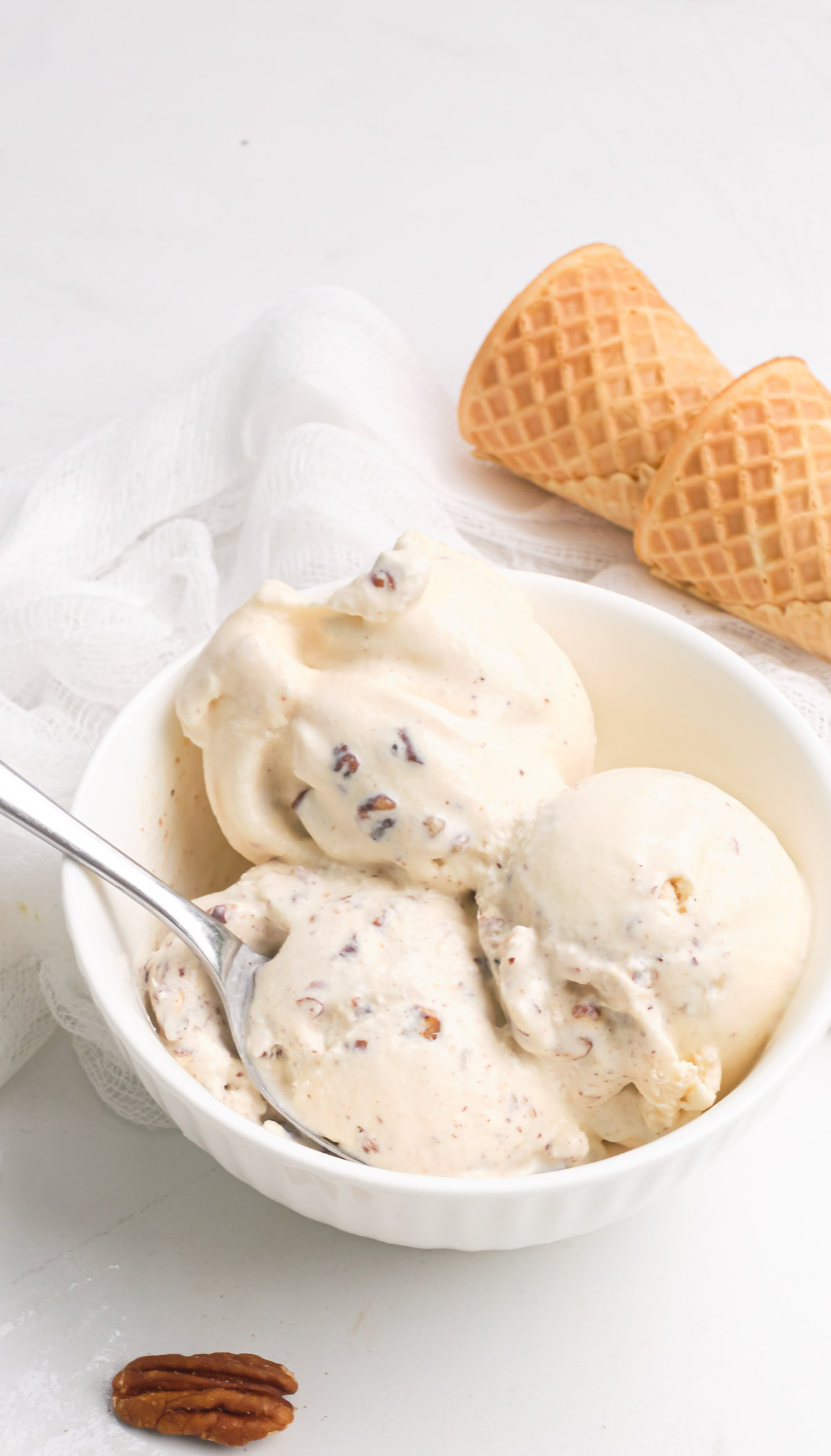 three scoops of the butter pecan ice cream in a white bowl with a spoon and two ice cream cones.