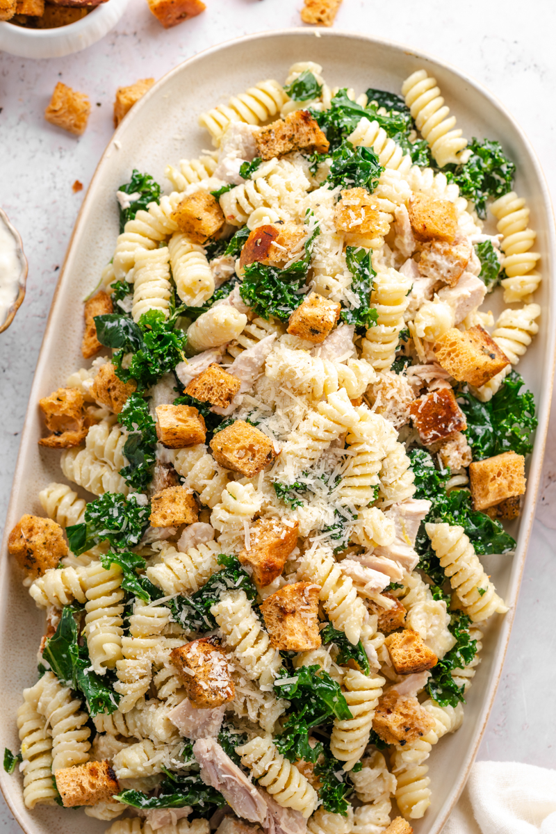 Large serving platter filled with a creamy pasta salad topped with crunchy croutons, kale, and shredded chicken.