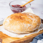 the air fryer bread on a wood cutting board set next to a blue checked kitchen towel and pot of jam.