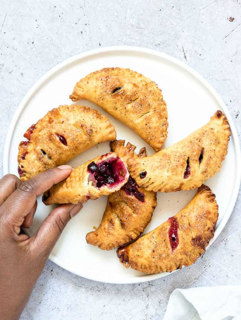 a hand holding one blueberry hand pie with a bite out of it in front of a white plate filled with air fryer blueberry hand pies