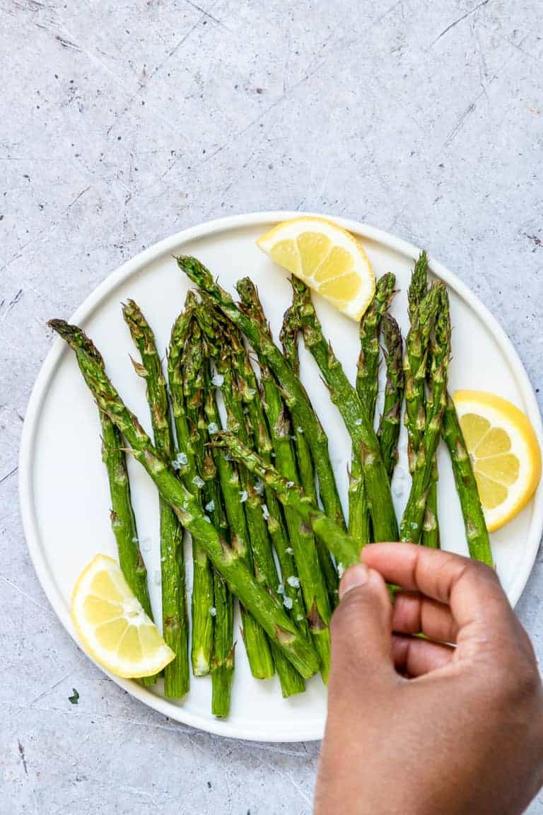 a hand removing one asparagus spear from a plate filled with air fryer asparagus and lemon wedges