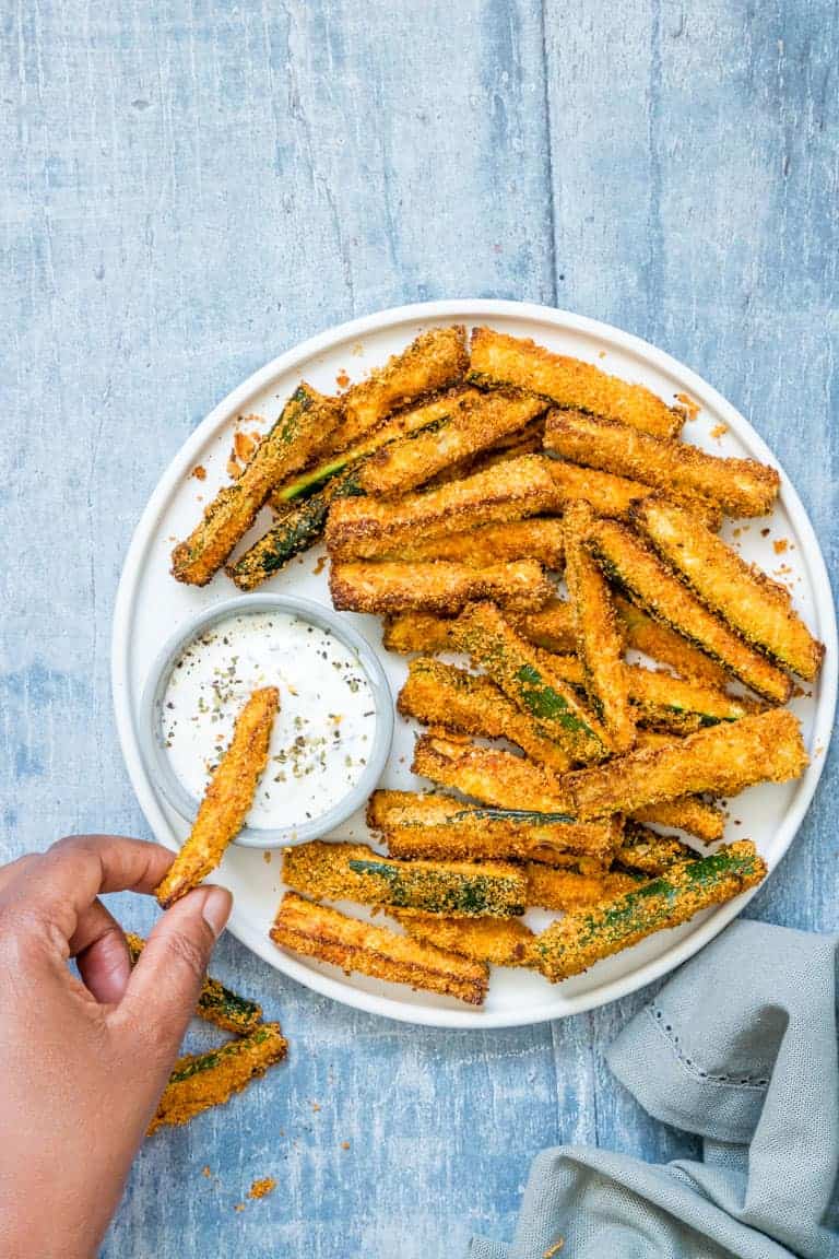 air fryer zucchini fries on a white plate with a hand dipping one of the zucchini fries into creamy herb yogurt dip