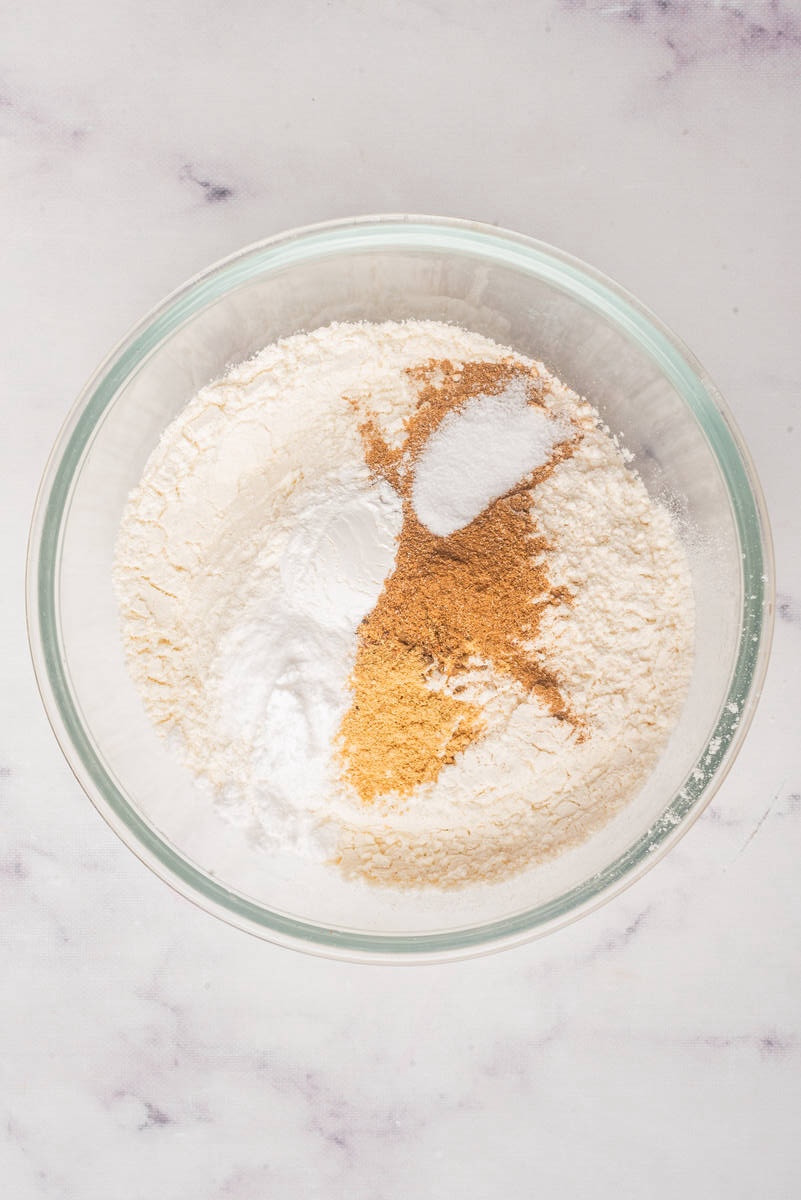 A glass bowl containing dry ingredients for baking, including flour, baking powder, baking soda, salt, and brown spices.
