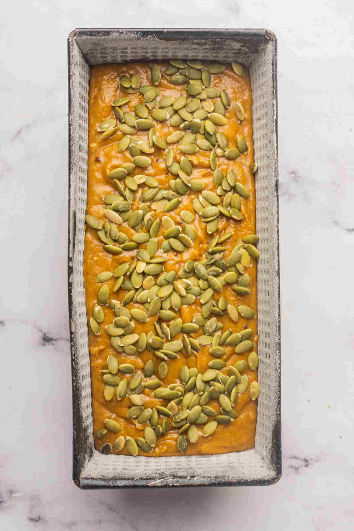 A loaf pan with batter topped with raw pumpkin seeds, ready to be baked.