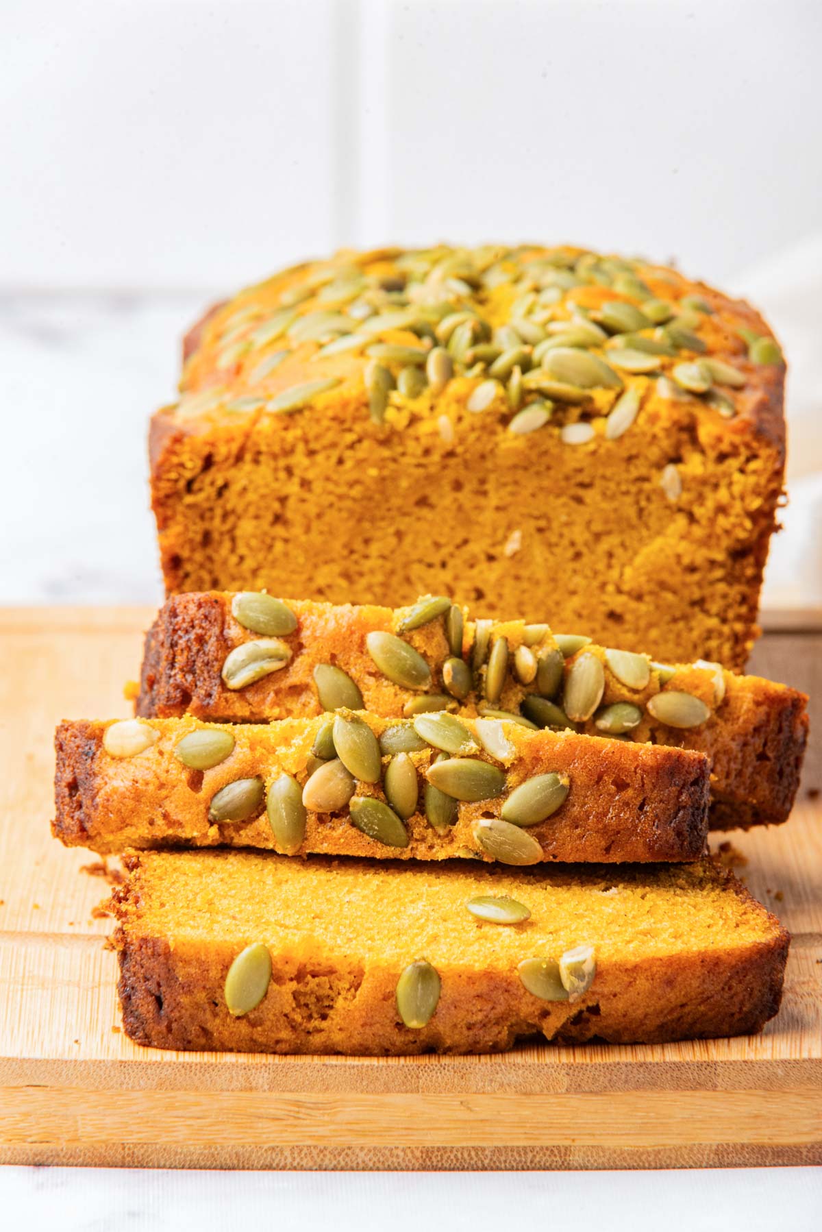 A loaf of pumpkin bread with a few slices cut and arranged in front, showcasing the interior texture and pumpkin seed topping.