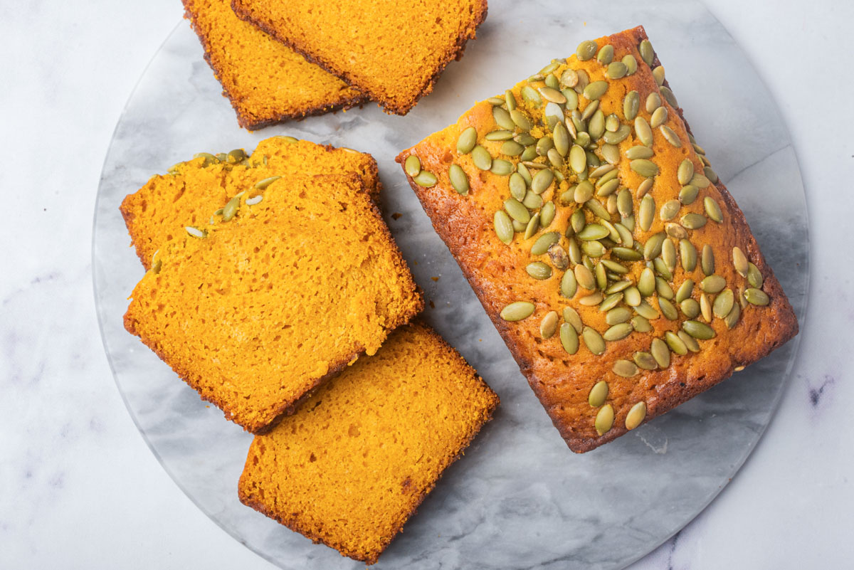 A top-down view of a loaf of pumpkin bread and several slices, displaying the rich orange color and seed topping on a marble surface.