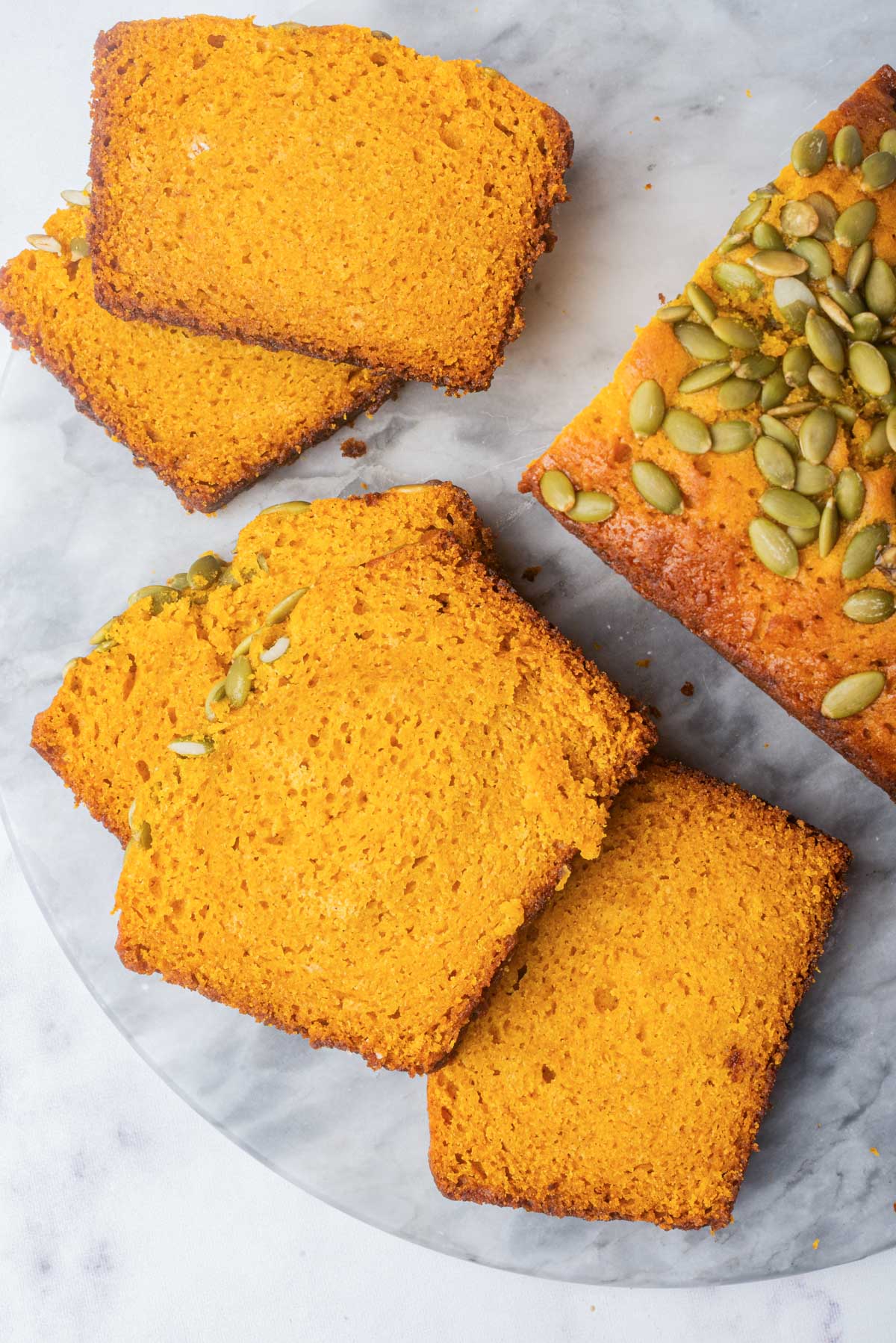 A top-down view of sliced pumpkin bread arranged neatly on a marble surface, highlighting the vibrant color and seed topping.