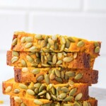A stack of sliced copycat Starbucks pumpkin bread with a hand picking up the top slice, emphasizing the texture and pumpkin seed topping.