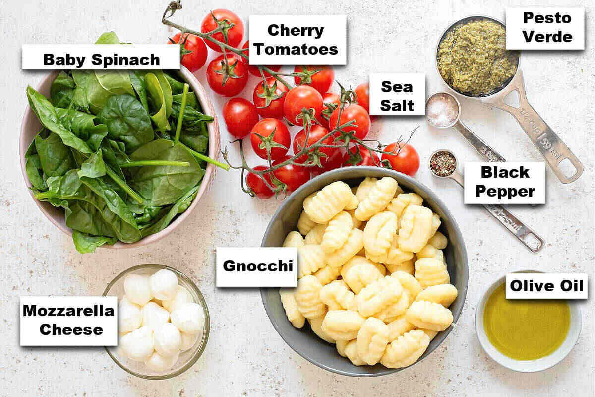 A display of ingredients for a sheet pan dinner, including baby spinach, cherry tomatoes, gnocchi, mozzarella cheese, pesto verde, sea salt, black pepper, and olive oil.