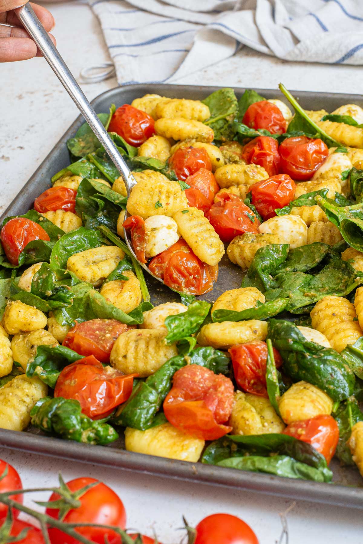 Gnocchi with cherry tomatoes and spinach on a sheet pan, showing a portion being served with a silver spoon.