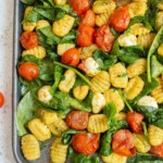 Baked gnocchi with cherry tomatoes and spinach on a sheet pan, showing a portion being served with a spoon.