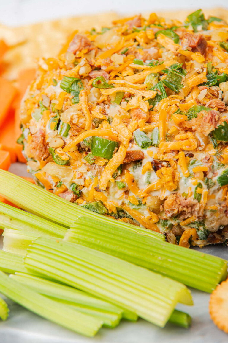 Close-up of cheese ball garnished with cheddar, bacon, and green onions, paired with fresh celery sticks.