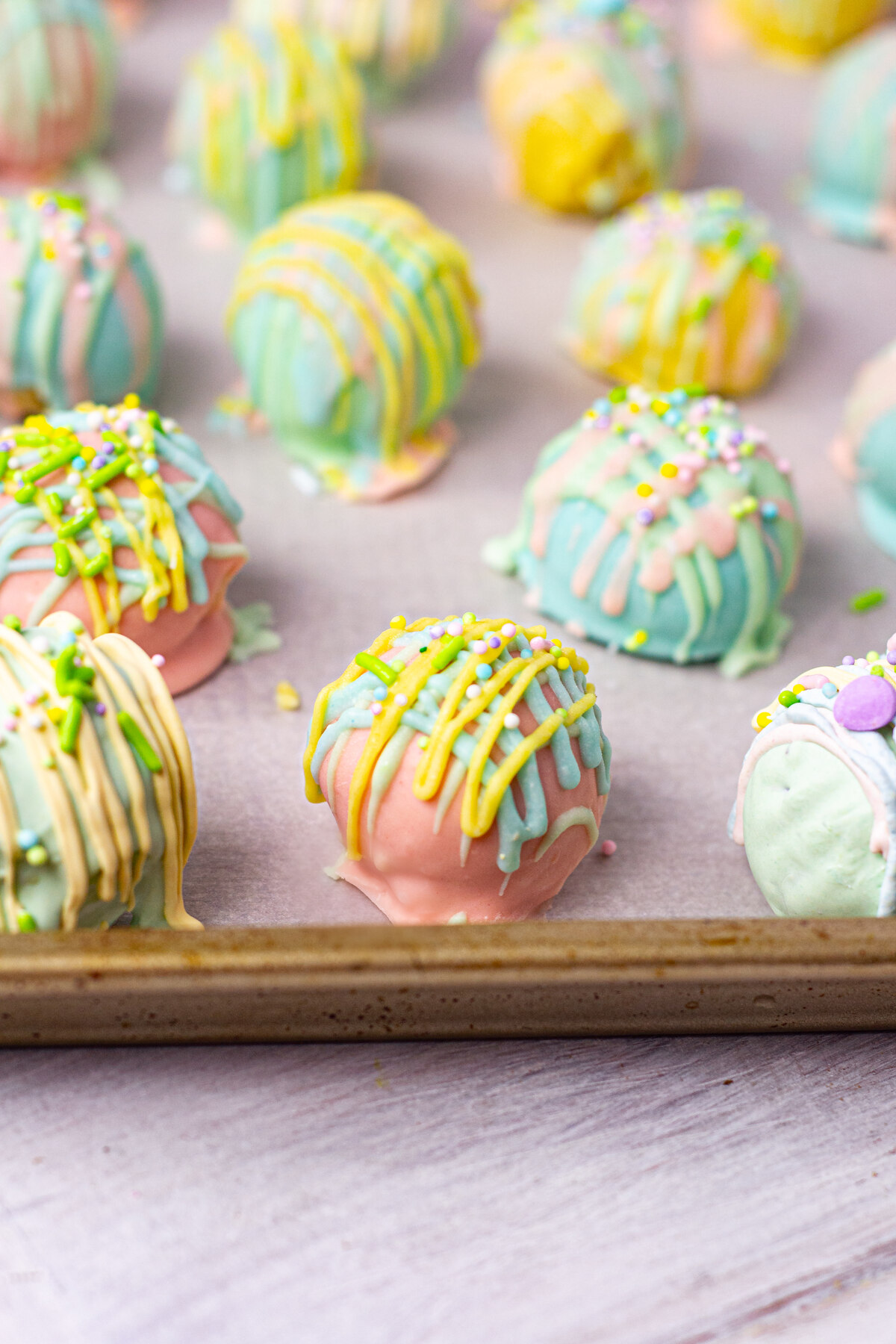 A close-up of pastel-colored Oreo truffles on a baking sheet. The truffles are coated in pink, blue, yellow, and green chocolate with drizzle decorations and tiny sprinkles.