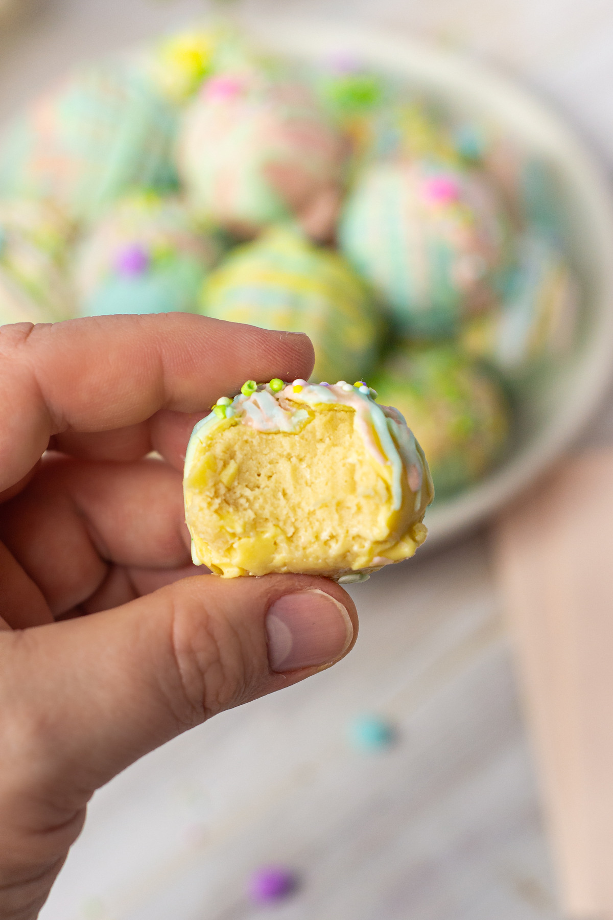 A hand holding a pastel-colored Oreo truffle with a bite taken out, revealing a creamy yellow filling. The background shows a plate filled with more decorated truffles.
