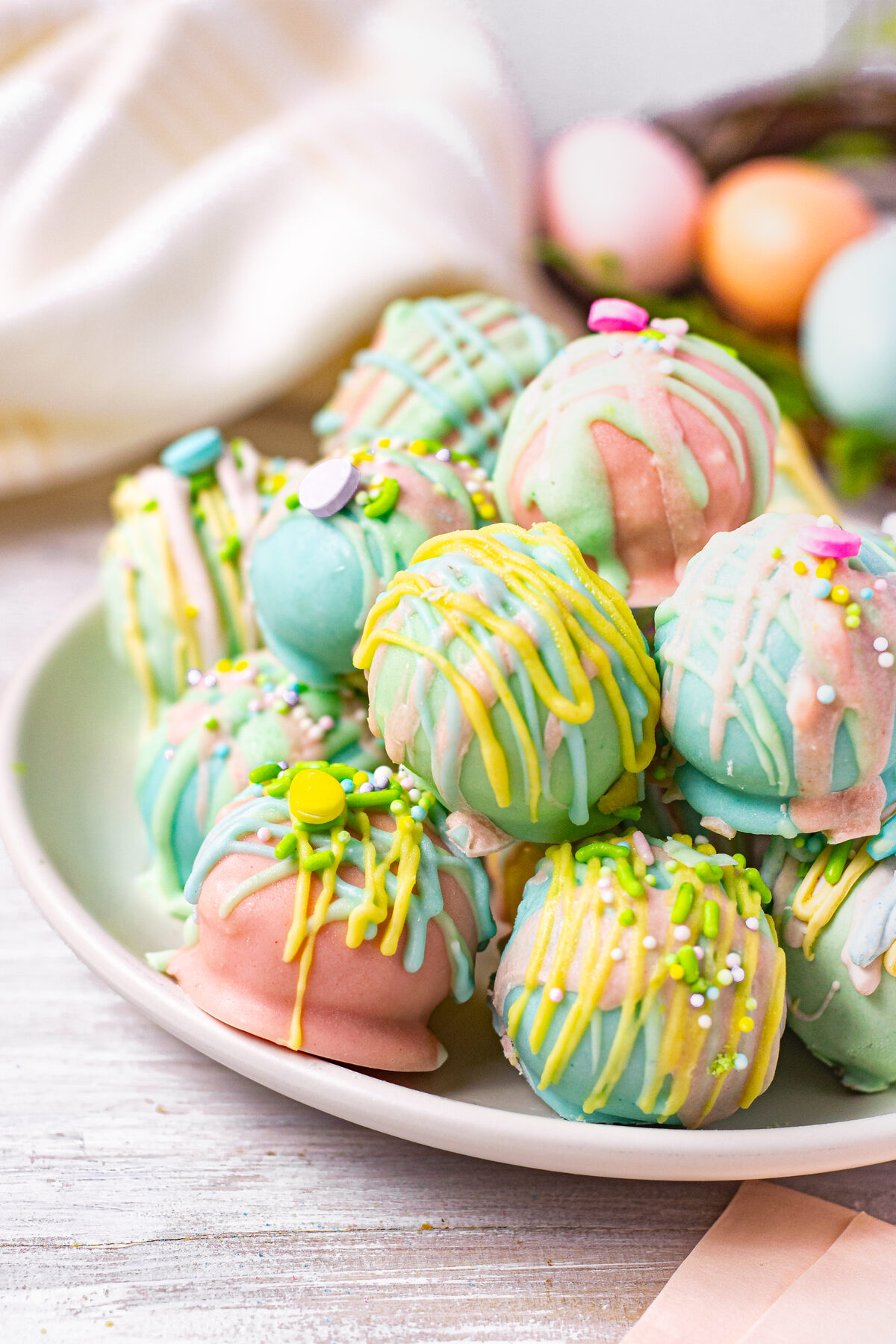 A plate of pastel-colored Oreo truffles with yellow, pink, and blue chocolate drizzle and colorful sprinkles. The soft background includes hints of Easter-themed decorations.