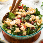 Tortellini pasta salad tossed with spinach, mozzarella pearls, kalamata olives, sun-dried tomatoes, and artichoke hearts in a large glass serving bowl.