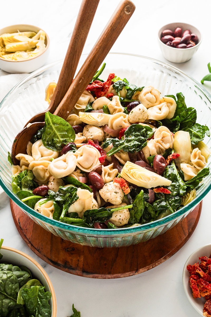 Tortellini pasta salad tossed with spinach, mozzarella pearls, kalamata olives, sun-dried tomatoes, and artichoke hearts in a large glass serving bowl.