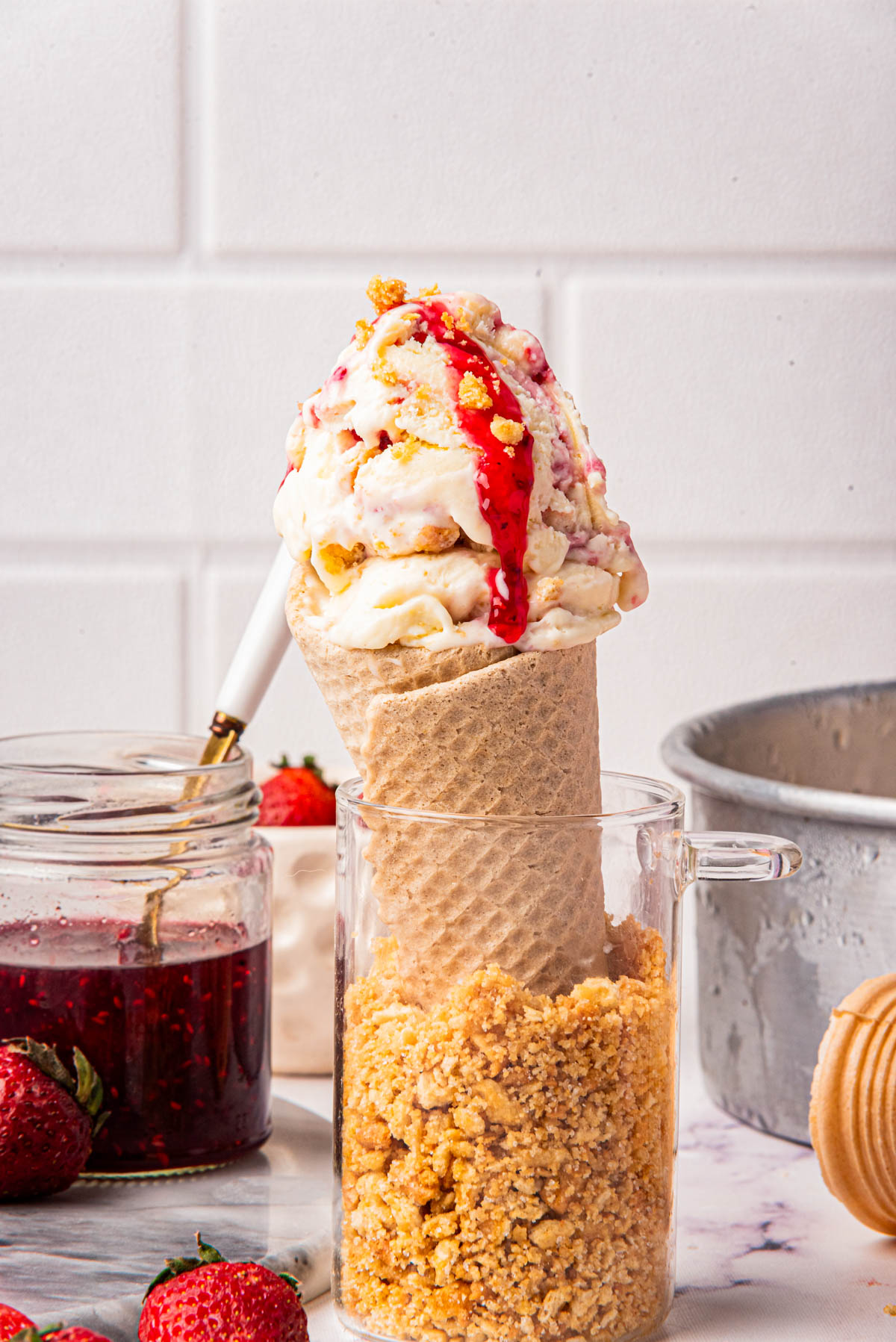 Close-up of strawberry ice cream served in a sugar cone with jam and graham cracker crumbs, with a background of strawberries and a striped napkin.