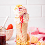 Close-up of strawberry cheesecake ice cream served in a sugar cone with jam and graham cracker crumbs, with a background of strawberries and a striped napkin.