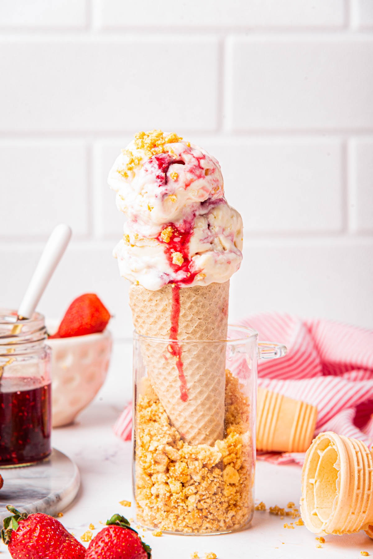 Close-up of strawberry cheesecake ice cream served in a sugar cone with jam and graham cracker crumbs, with a background of strawberries and a striped napkin.