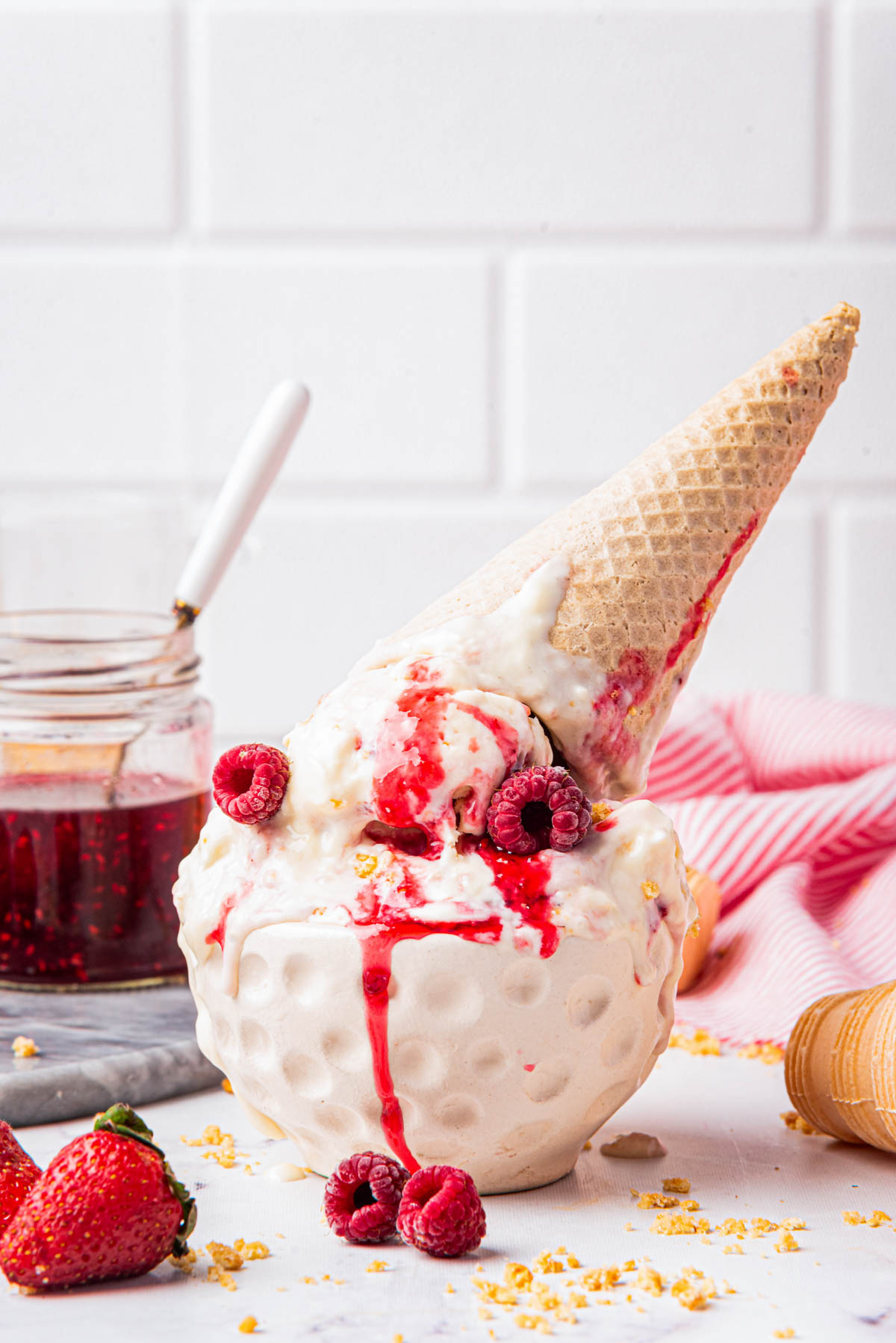Ice cream overflowing from a cone, topped with raspberries and graham cracker crumbs, with strawberries and a striped napkin nearby.