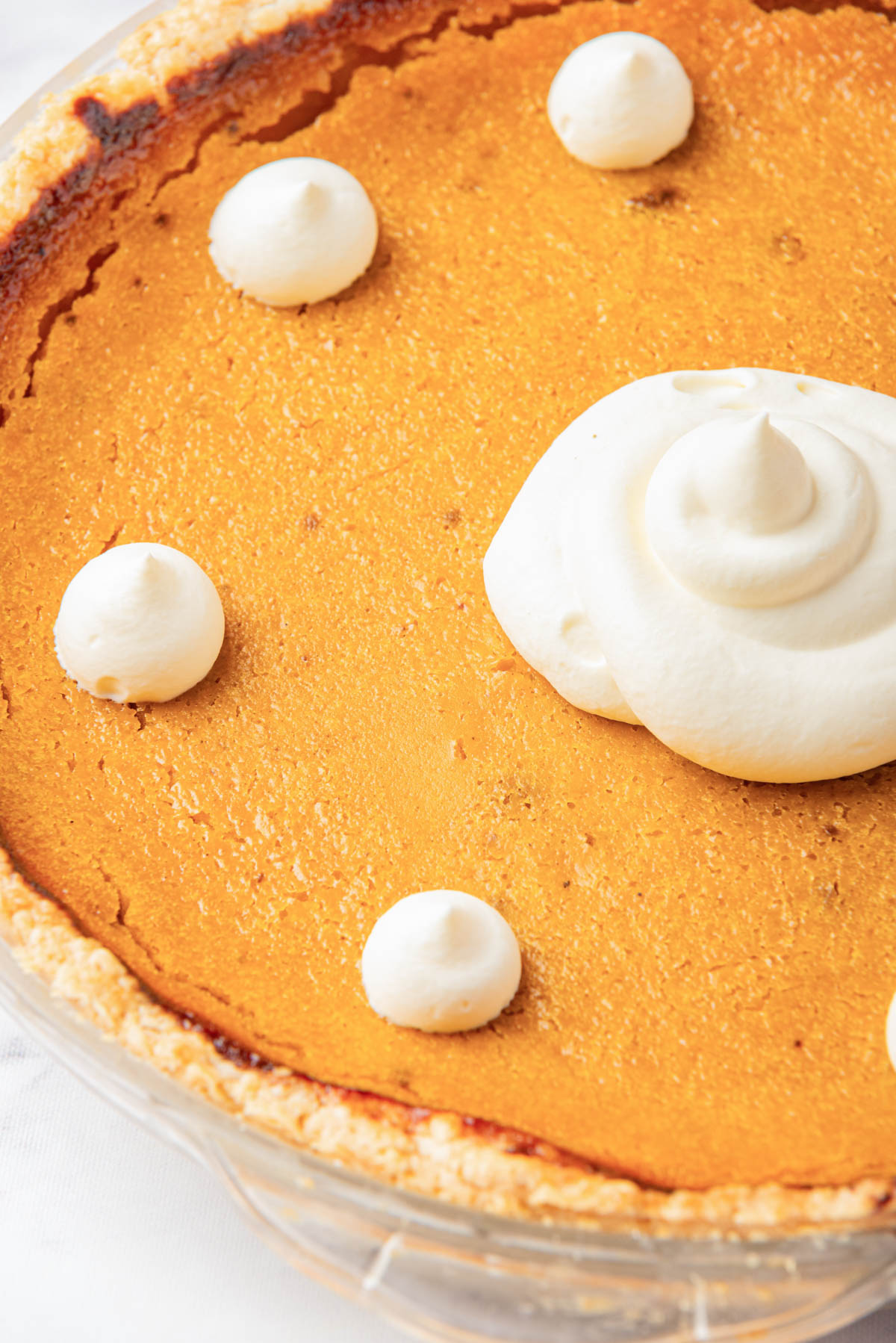 An overhead view of a whole pumpkin pie, centered with a large swirl of whipped cream and surrounded by smaller dollops, with a golden-brown crust.