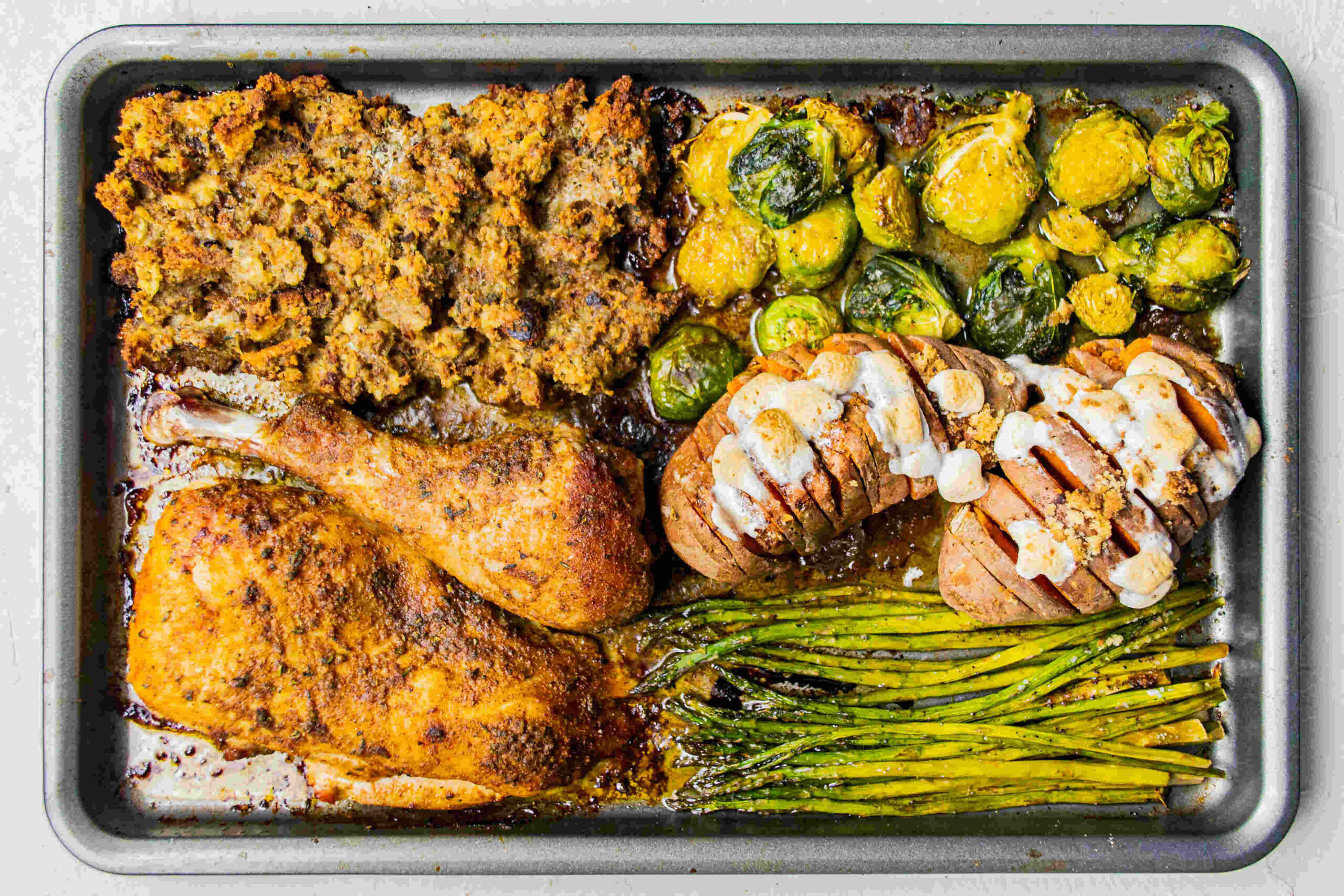 Overhead shot of a full sheet pan loaded with a complete turkey dinner—roasted turkey, golden stuffing, sweet potatoes with melted marshmallows, Brussels sprouts, and asparagus, all cooked together.