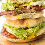 Close-up of a stacked breakfast sandwich with English muffins, smashed avocado, crispy bacon, a fried egg, and a sausage patty, with yolk dripping down the sides.