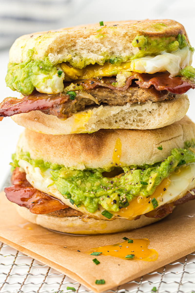Close-up of a stacked breakfast sandwich with English muffins, smashed avocado, crispy bacon, a fried egg, and a sausage patty, with yolk dripping down the sides.