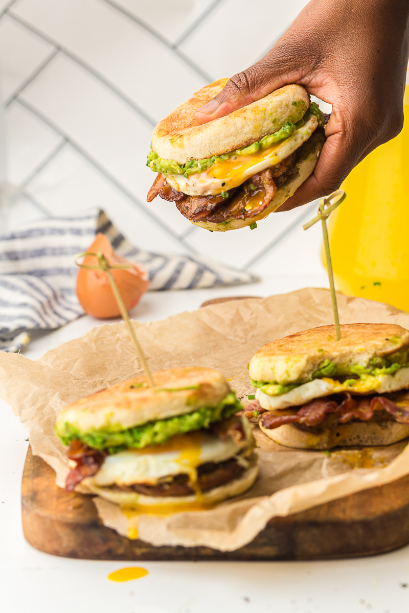 Three breakfast sandwiches on a wooden board, one being held by hand, each layered with avocado, egg, bacon, and sausage on toasted English muffins.
