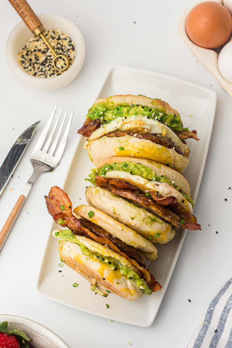 Four breakfast sandwiches lined up on a white platter, filled with smashed avocado, egg, bacon, and sausage on toasted English muffins, sprinkled with herbs and seeds.