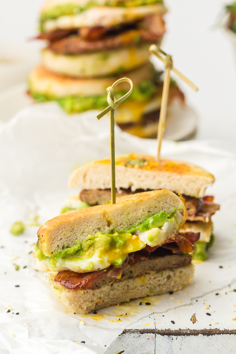 Cross-section of a breakfast sandwich secured with a skewer, showing layers of smashed avocado, crispy bacon, fried egg, and sausage on an English muffin, with egg yolk oozing out.