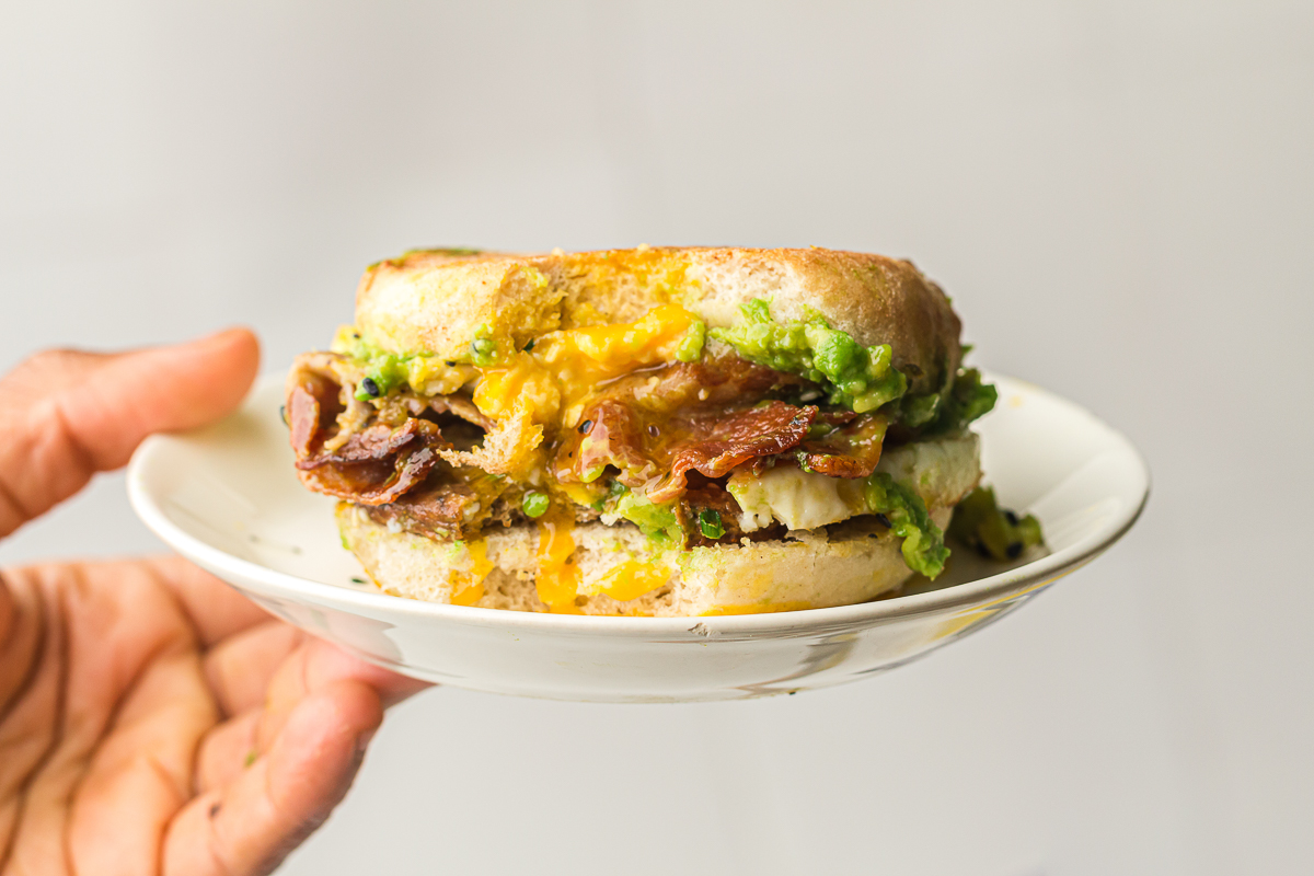 Hand holding a breakfast sandwich on a white plate, filled with crispy bacon, avocado spread, egg, and melted cheese on an English muffin.