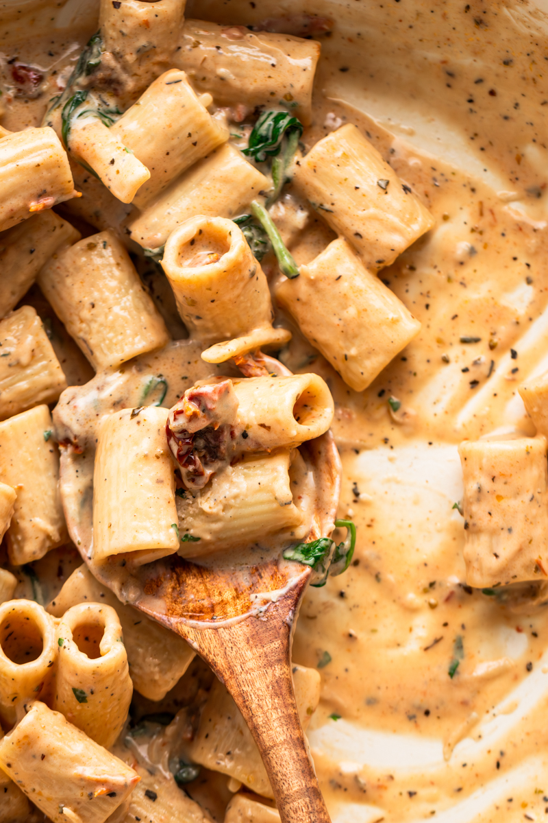 Close-up of creamy pasta mixed with tender rigatoni, spinach, and herbs in a white pan with a wooden spoon.